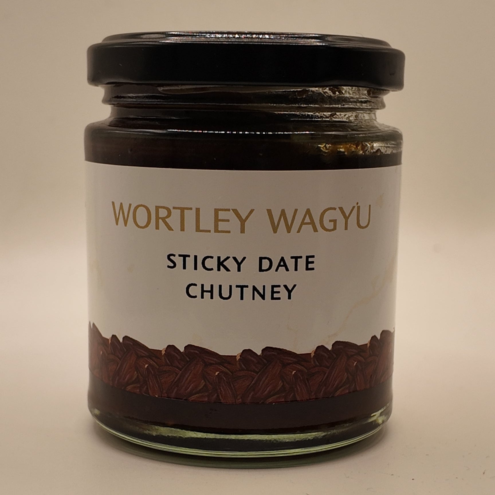 A glass jar of Wortley Wagyu Sticky Date Chutney with a black lid and a white label, set against a plain beige background.
