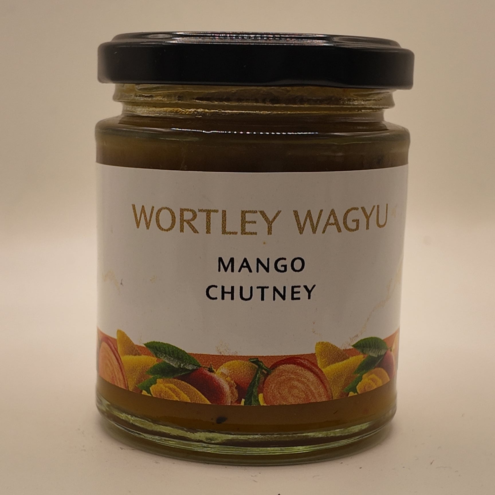 A glass jar of Wortley Wagyu Mango Chutney with a black lid and a white label featuring orange and yellow fruit illustrations, set against a plain light background.