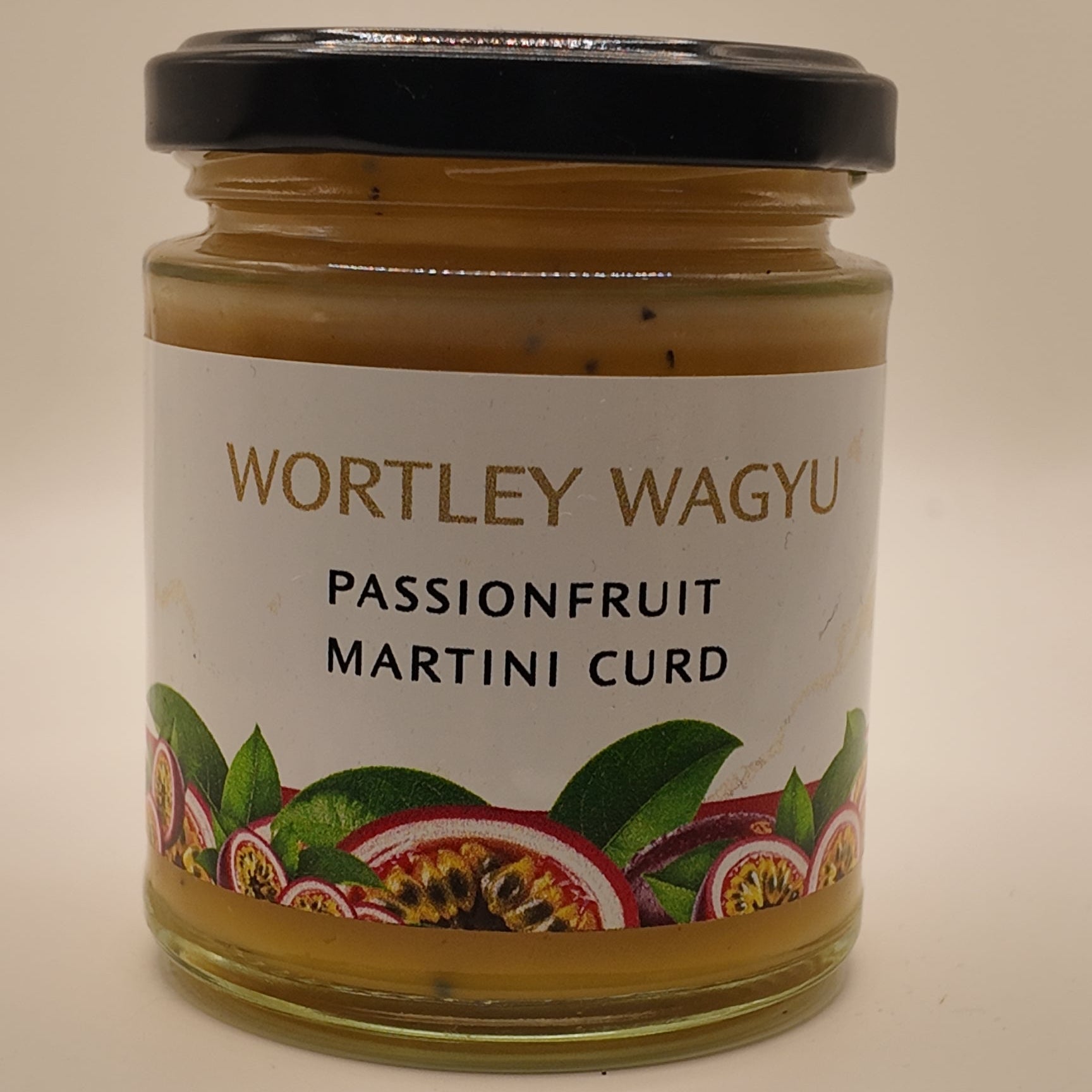 A glass jar of Wortley Wagyu Passionfruit Martini Curd with a black lid and a white label featuring passionfruit illustrations, set against a plain beige background.