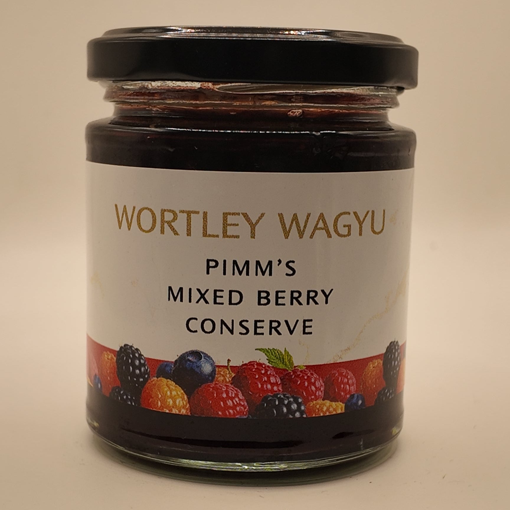 A glass jar of Wortley Wagyu Pimm's Mixed Berry Conserve with a black lid and a white label featuring colorful berries, set against a plain beige background.